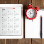 tablet with calendar for 2021, alarm clock and a spring notebook on a wooden background flat lay