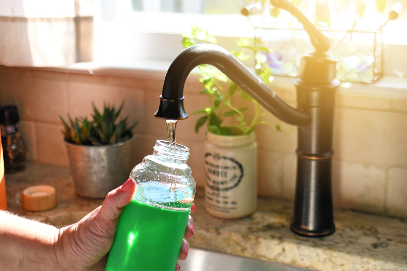 Woman filling reusable eco-friendly glass water bottle in sink, environmentally friendly container