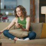 Relaxed girl eating snacks at cozy couch. Carefree woman browsing mobile phone