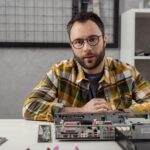 man looking at camera against broken computer on table