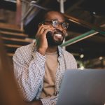 Happy male IT professional in spectacles using laptop and smartphone for call