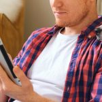 man swipes through a tablet, while his intrigued dog leans in, resting its head on the man’s